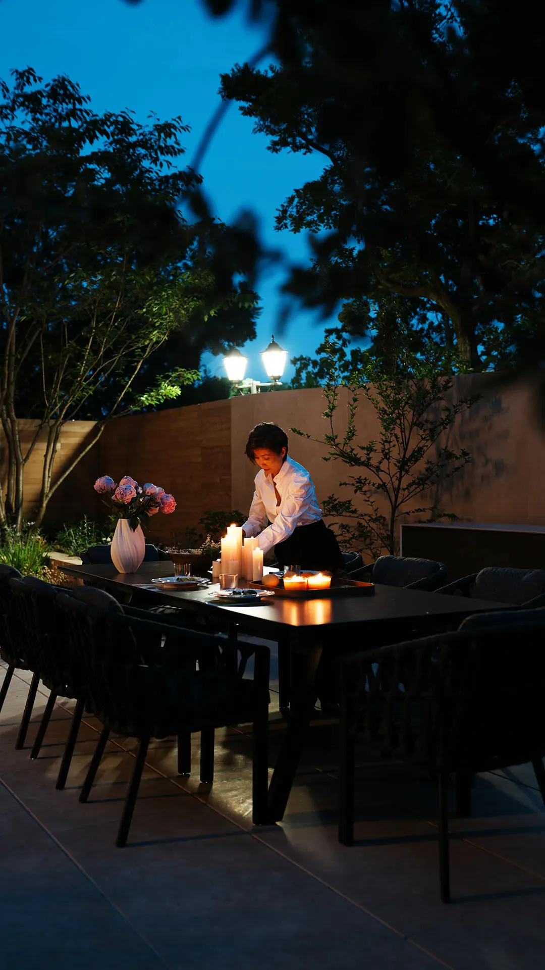 Woman setting a long outdoor dining table with lit candles, flower vase, and plates in a twilight garden with trees and wall lighting.