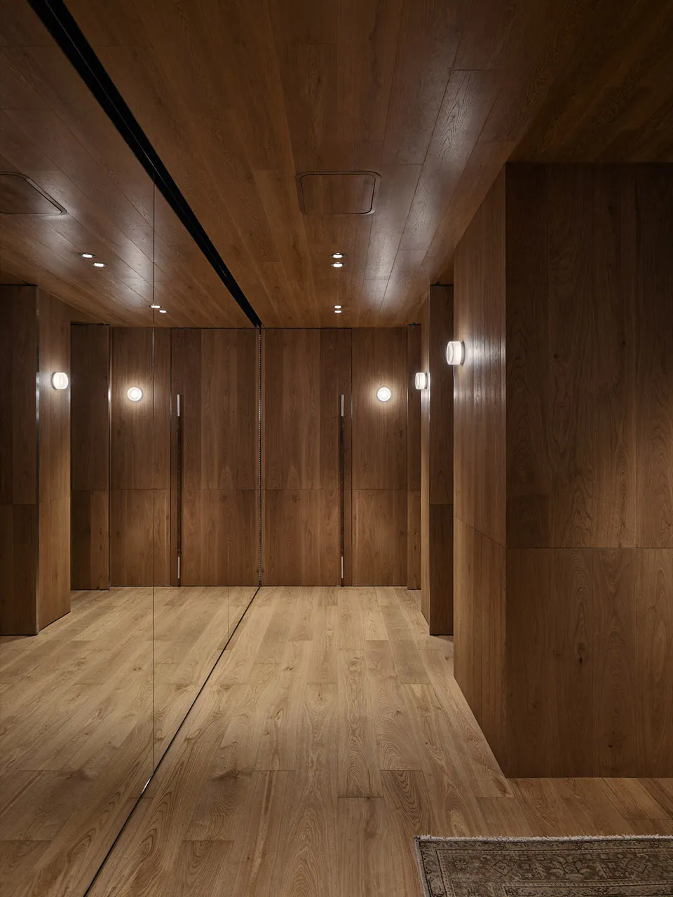 Modern walk-in closet interior featuring rich walnut wood paneling, full-height mirrored closet doors, recessed ceiling lights, and warm wall sconces.