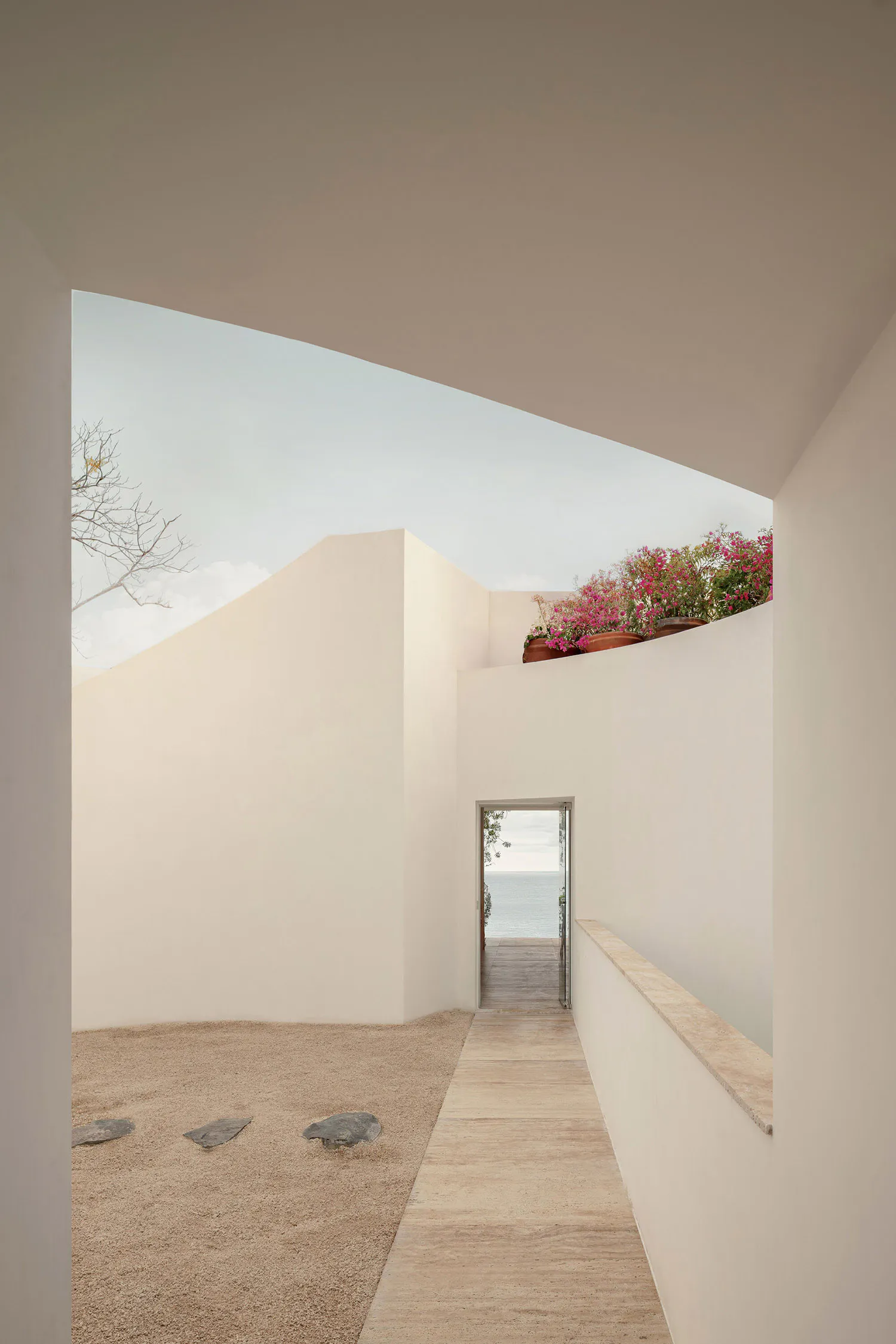 Modern minimalist courtyard with sand ground, stepping stones, a terrazzo walkway, white plastered walls, bougainvillea on a rooftop garden, and a view of the sea through an open doorway.