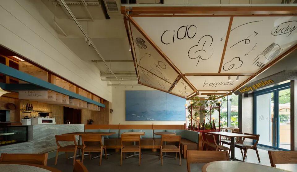 Modern cafe interior with wooden furniture, marble counter, built-in booth seating, and a large suspended ceiling structure covered in black hand-drawn words and doodles including 'ciao', 'happiness', 'lucky garden', and 'a table full of joy'.