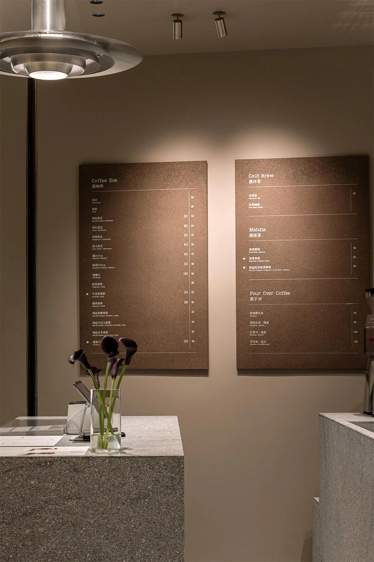 Modern minimalist cafe interior with two large brown menu boards displaying coffee and matcha selections, a speckled terrazzo counter in the foreground holding a glass vase with dark purple calla lilies, and contemporary pendant and spot lighting.