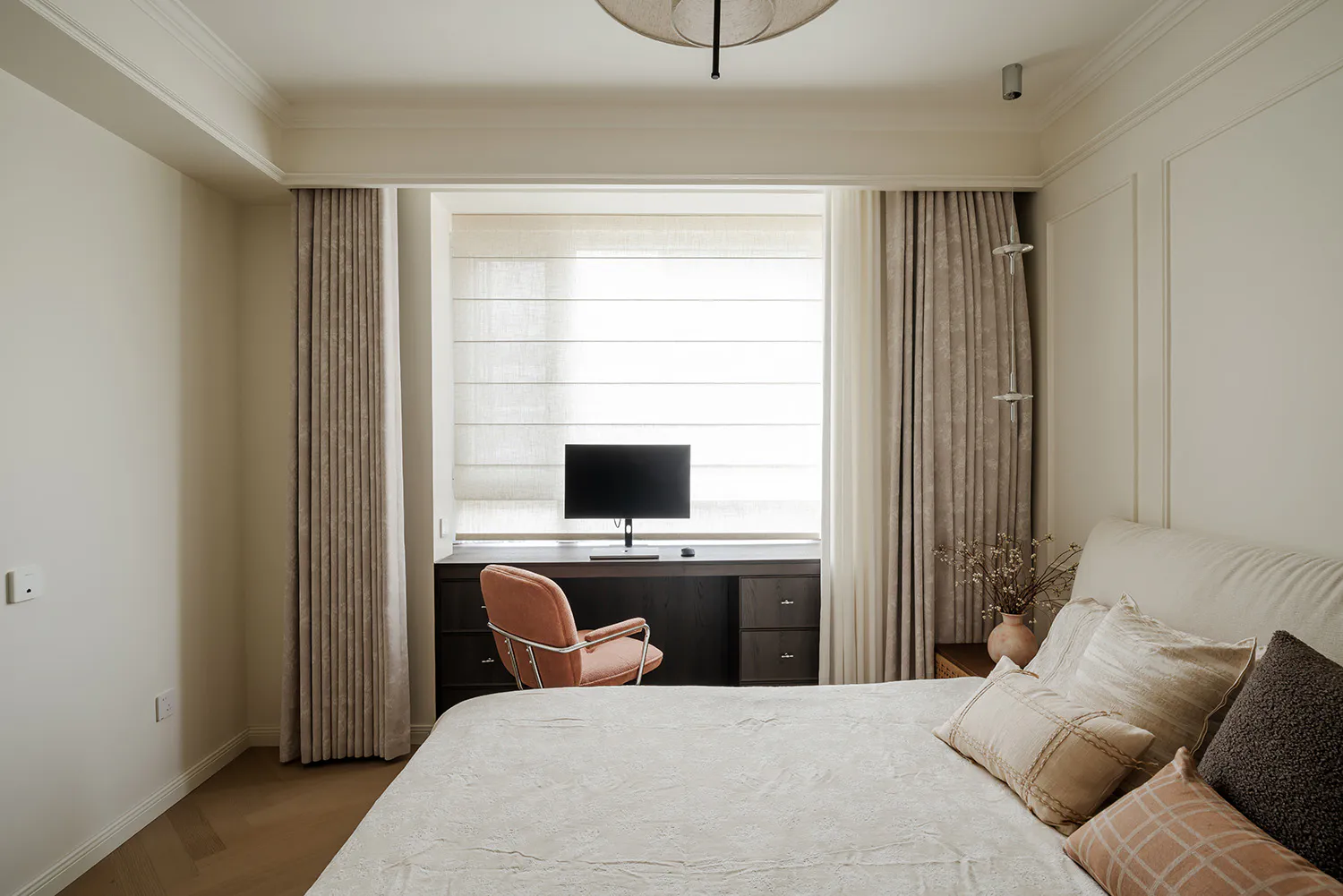 Modern neutral bedroom featuring a large bed with layered beige and brown pillows, a built-in dark wood desk under the window with a coral velvet chair, and soft linen curtains.