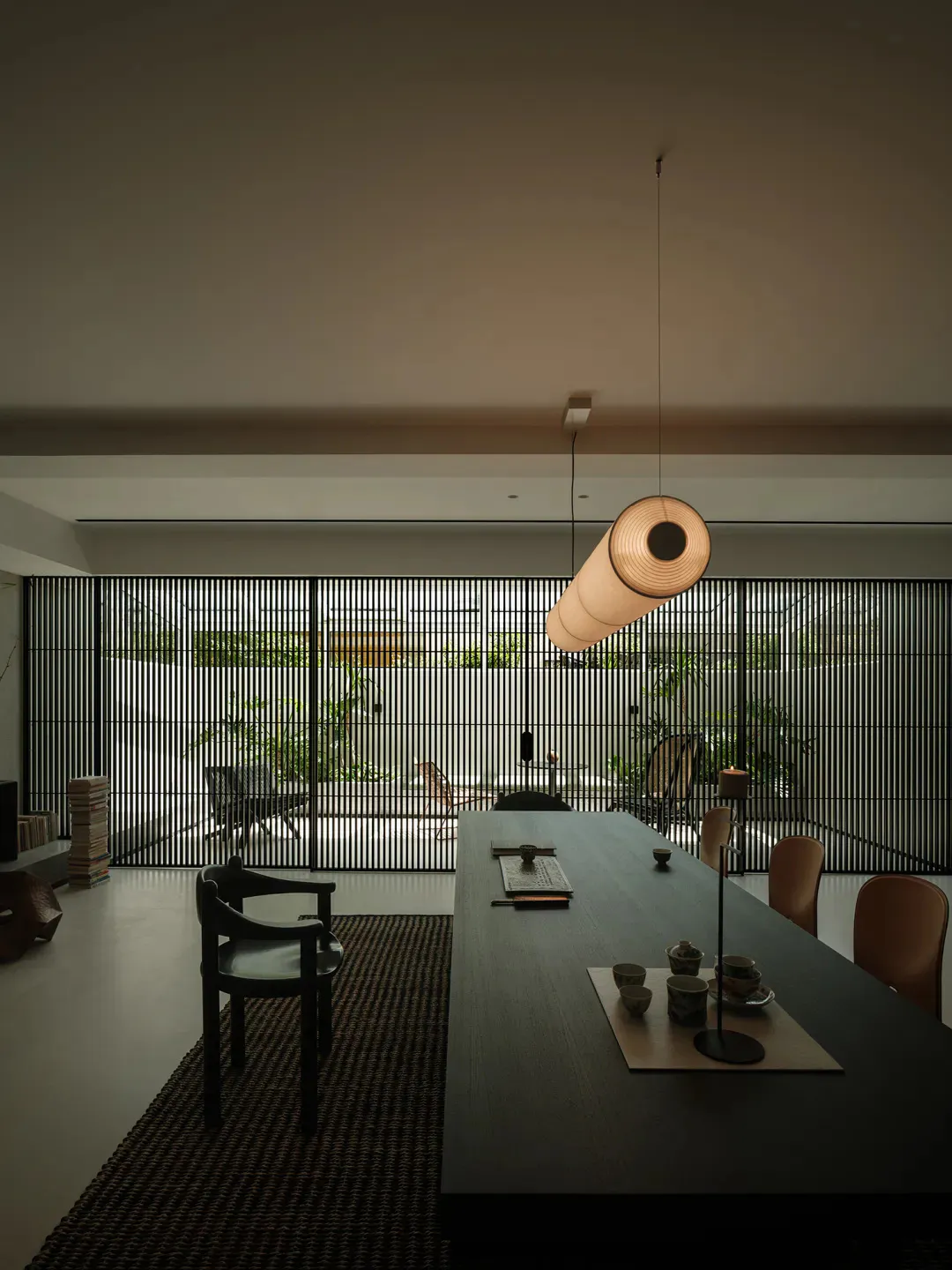 Modern minimalist dining room featuring a long dark wood table, black wooden chairs, a large cylindrical fabric pendant light, and a full-width black slatted screen opening to a lush green courtyard.