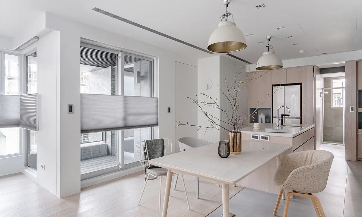 Modern minimalist dining room with white walls, light wood flooring, a rectangular dining table, upholstered chairs, and an open kitchen with light wood cabinetry in the background.