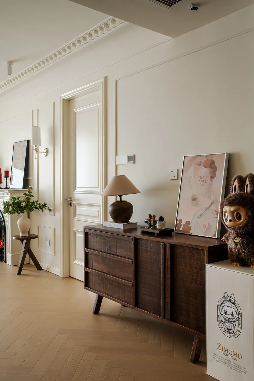 Modern classic hallway with off-white walls, crown molding, dark walnut wooden sideboard, table lamp, framed art, and Labubu doll on a white storage box.
