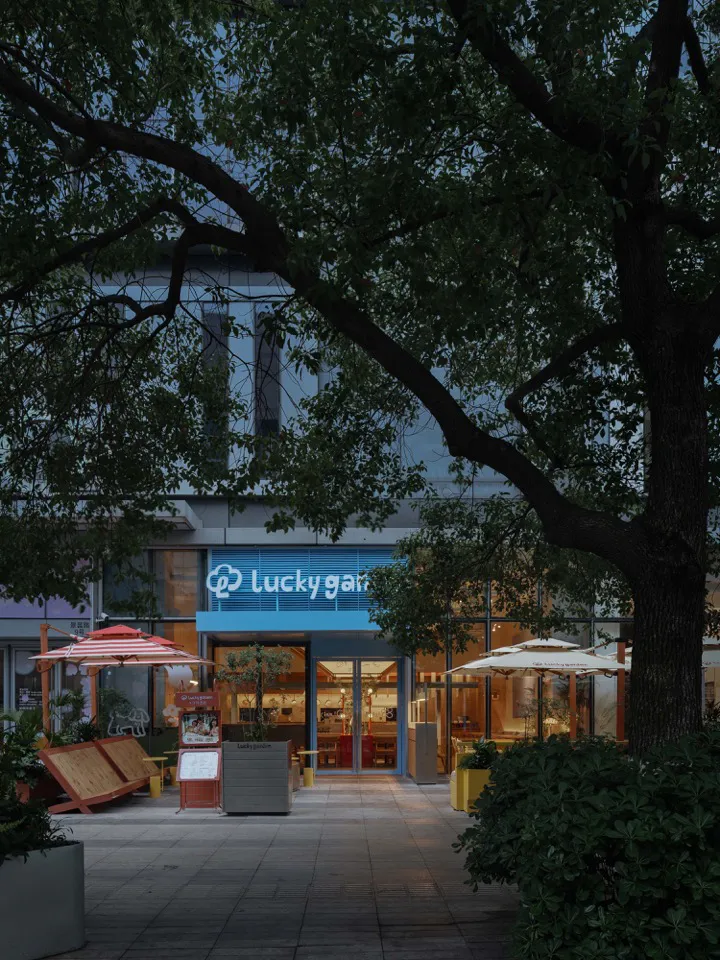 現代咖啡廳店面,藍色發光招牌寫著Lucky Garden,玻璃入口大門、戶外遮陽傘,以及黃昏時的茂密樹木。