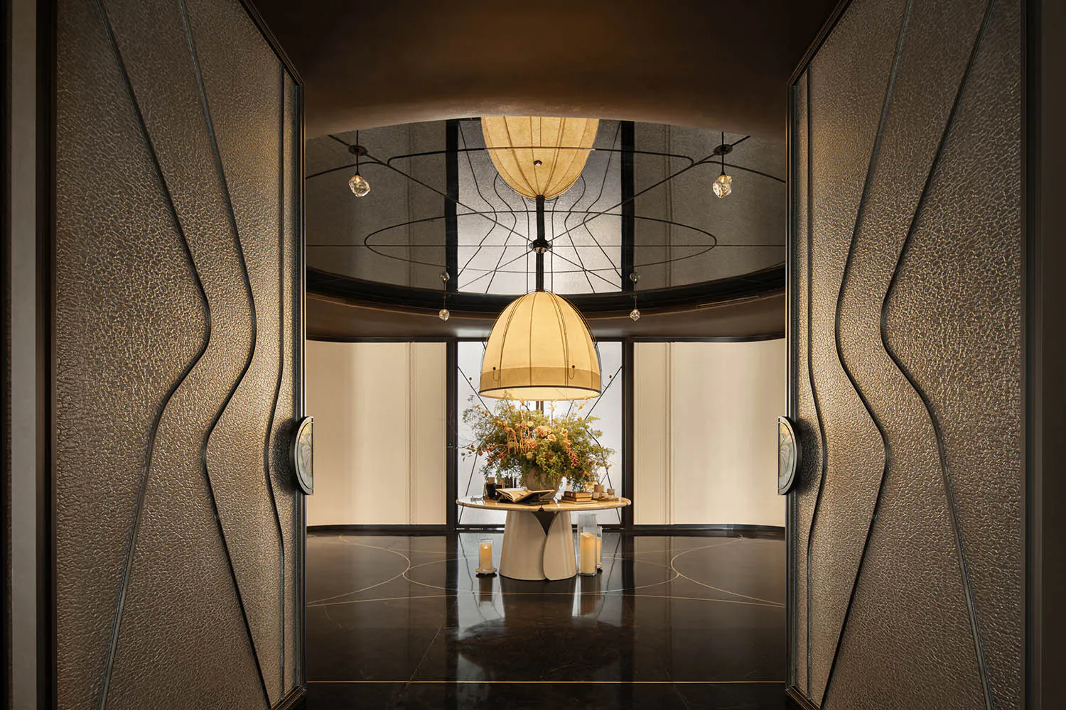 View through open textured leather doors into a luxurious circular foyer featuring a large floral arrangement on a sculptural table, dramatic layered pendant lights, and a glossy dark marble floor with gold inlay.