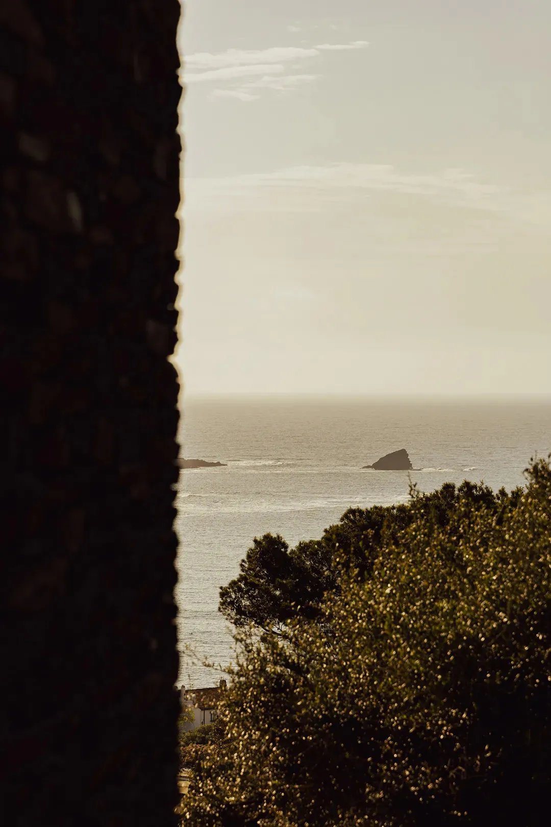 View of the ocean and a rocky island through a rough stone window or doorway, with green trees and a white building visible below.