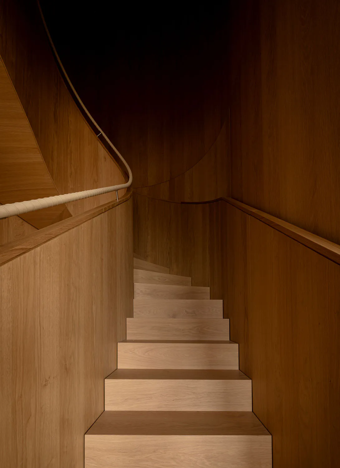 View looking down a curved wooden staircase made of light oak with a thick rope handrail mounted on a wooden wall.
