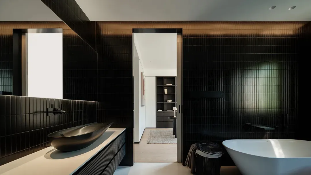 Modern luxury bathroom with black ribbed tiles, a freestanding white bathtub, bronze vessel sink on a white countertop, and a view into a walk-in closet.