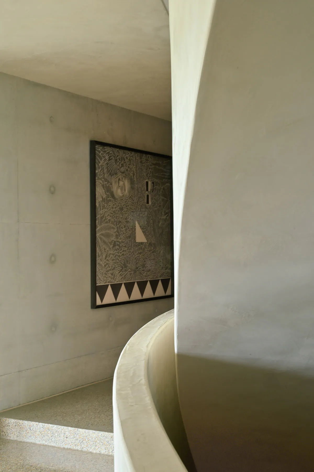 View of a curved concrete staircase and wall featuring a large framed artwork with intricate tropical etching and geometric border patterns against exposed concrete walls.