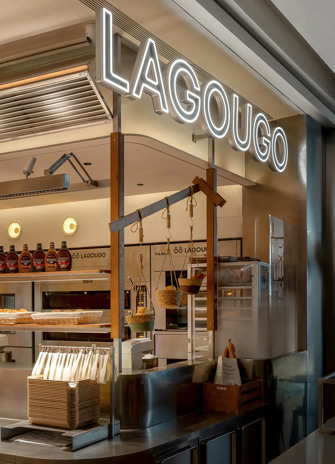Lagougo 現代麵包店吧台,大型白色霓虹招牌、木質結構橫梁、懸掛藤編麵包籃、不鏽鋼檯面與溫暖照明。