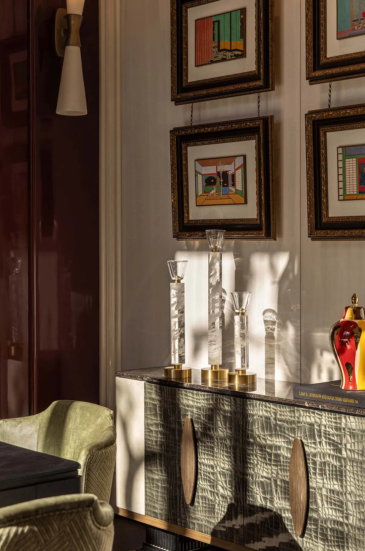 A luxurious dining room corner featuring a crocodile-embossed green sideboard topped with mother-of-pearl candle holders, colorful framed art on the wall, a modern wall sconce, and green velvet dining chairs.