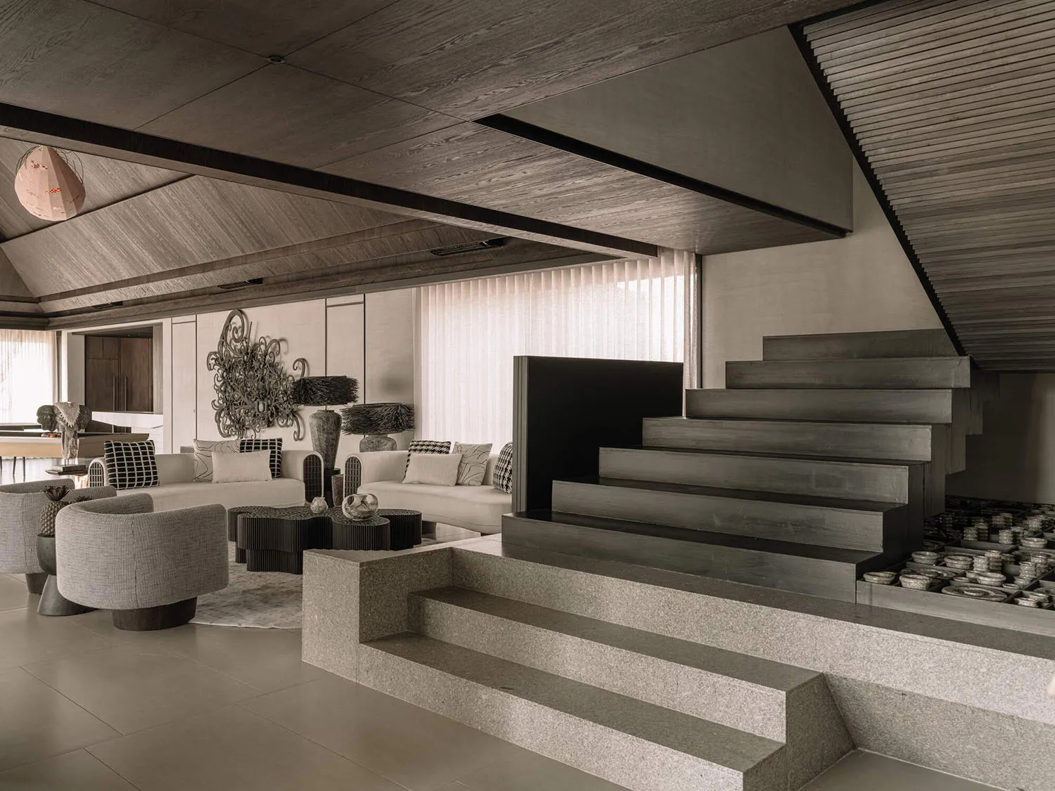 Modern split-level living room featuring a massive concrete staircase, neutral-toned sofas, textured grey armchairs, and dramatic wooden ceiling details.