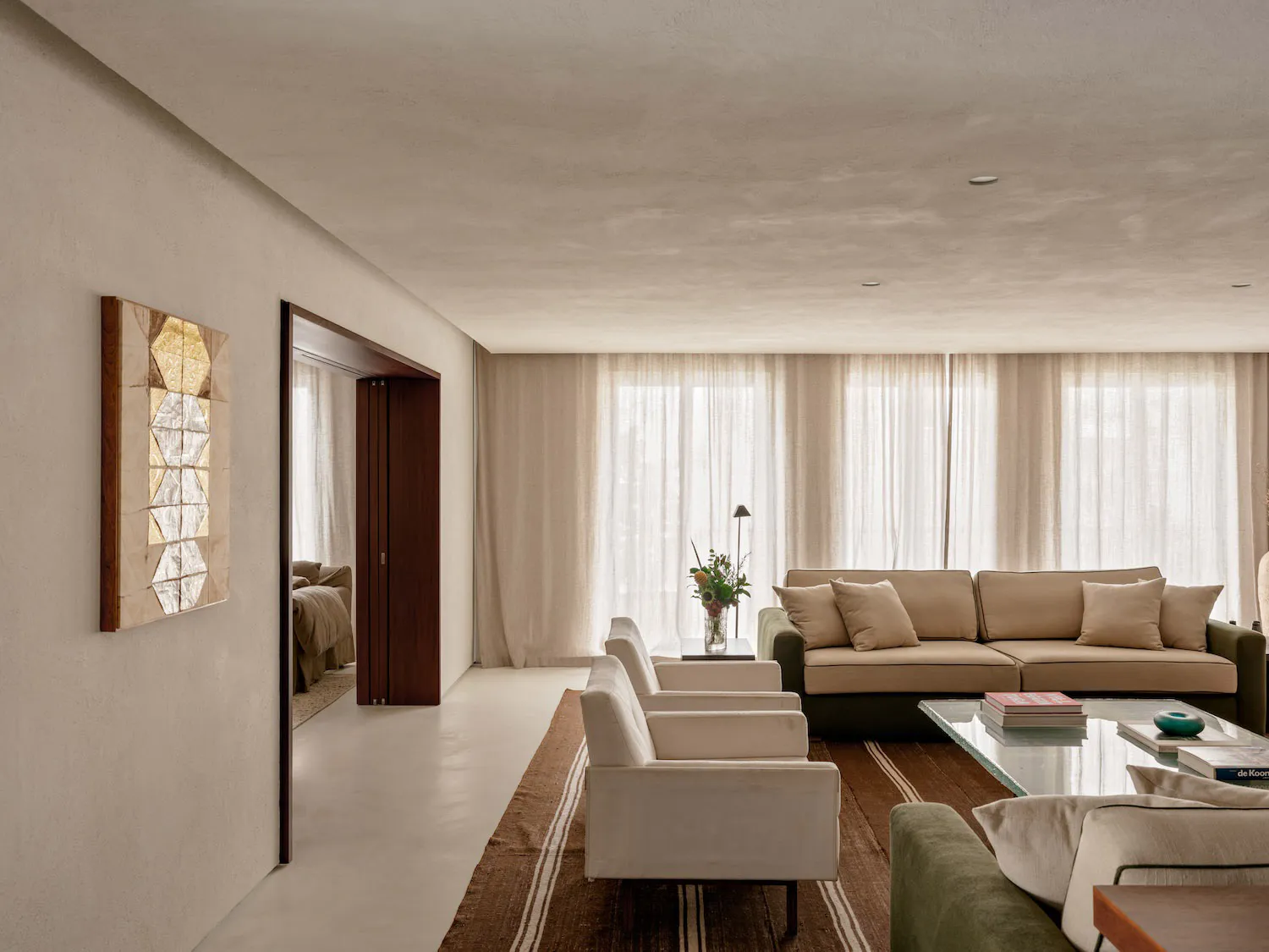 Modern minimalist living room with off-white plaster walls, beige sectional sofa, white armchairs, dark wood door, large windows with sheer curtains, and a striped brown rug.