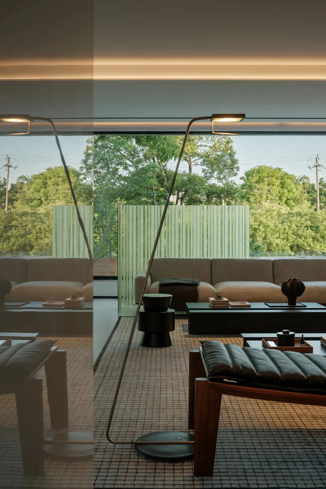 Modern minimalist living room featuring tatami mat flooring, a low beige sectional sofa, dark wood lounge chair, black side tables, and large glass windows overlooking green trees and a bamboo screen.