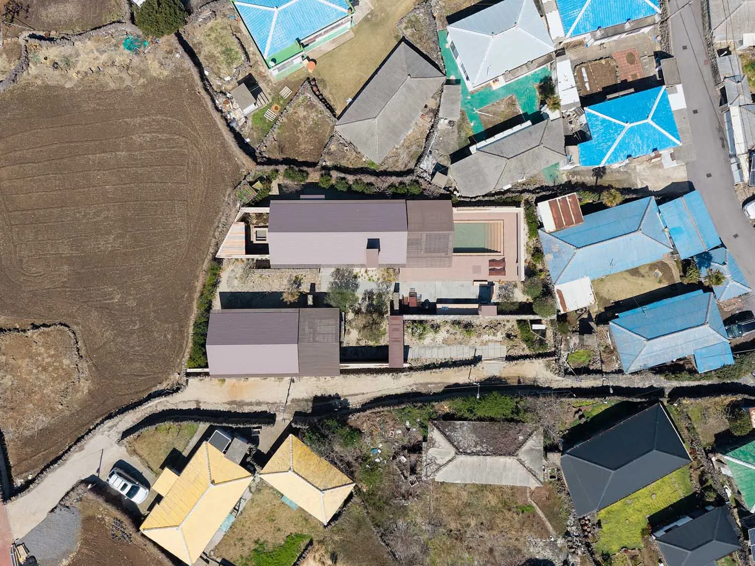 Aerial photograph of a modern single-story villa with a purple metal roof, rectangular swimming pool, and landscaped courtyard, situated among traditional houses with blue and yellow roofs in a rural Taiwanese setting.