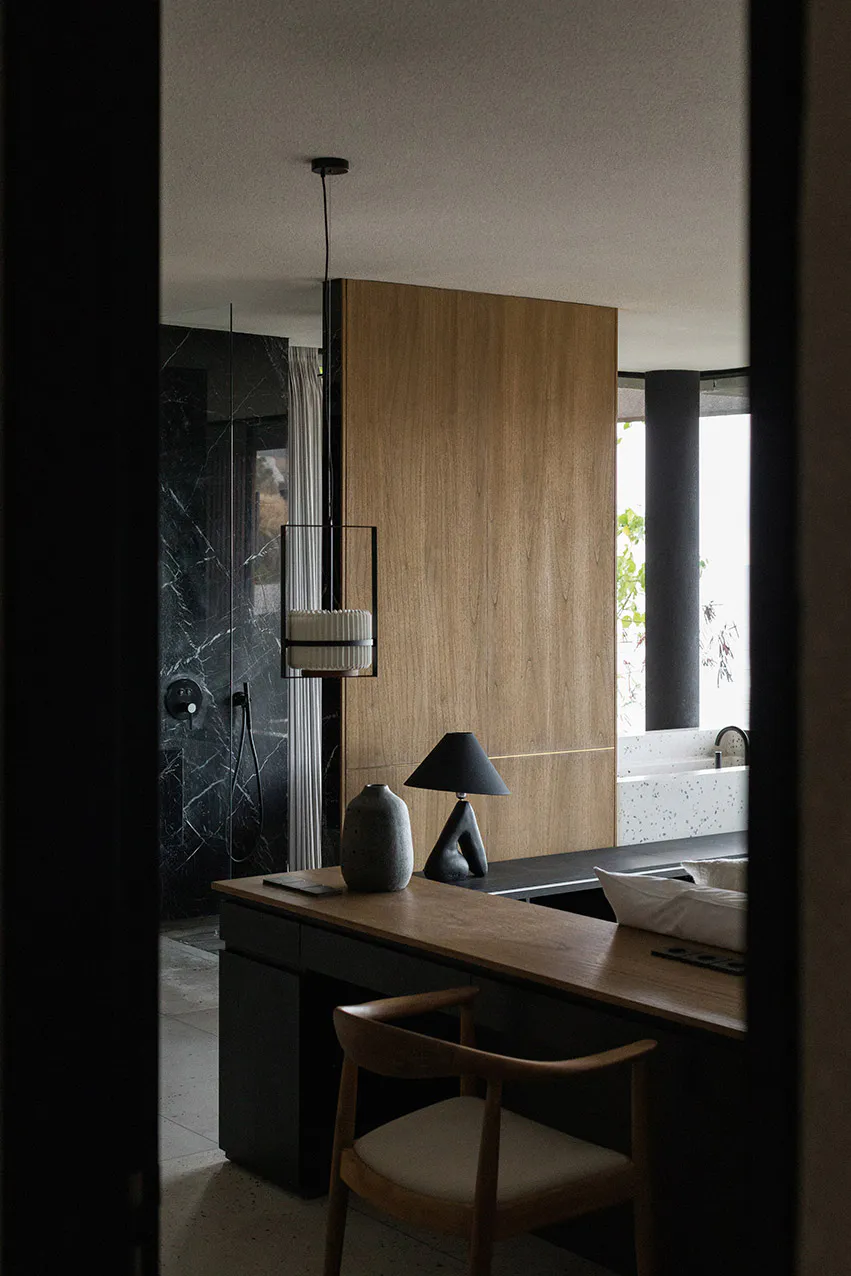 Modern bathroom interior viewed through a doorway, featuring a wooden vanity desk with a sculptural black lamp and ceramic vase, a wooden chair, a large warm wood panel wall, a black marble shower area, and a white terrazzo bathtub by a window.