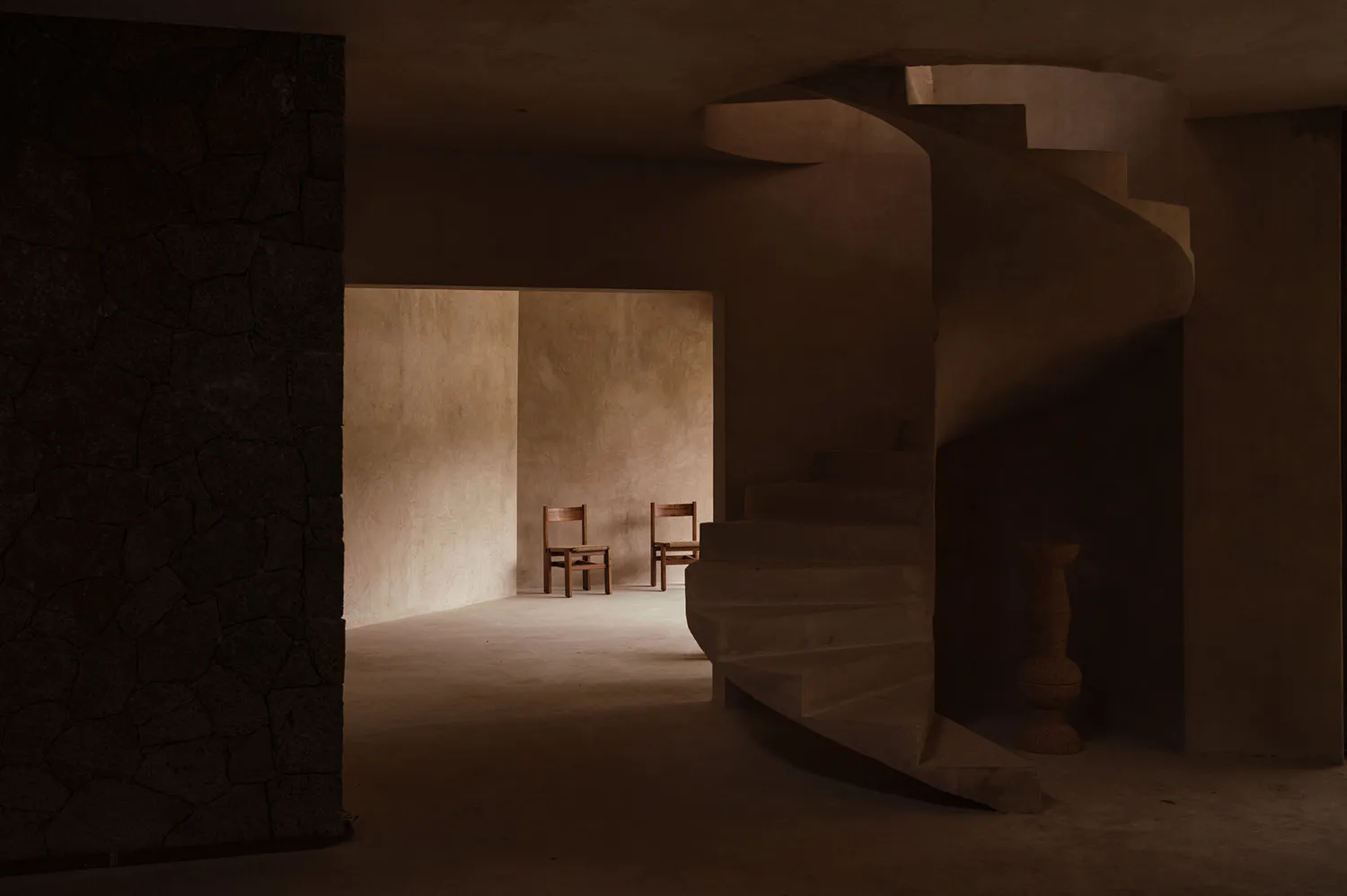 View of a sculptural concrete spiral staircase next to a dark volcanic stone wall in a dimly lit minimalist interior with wooden chairs visible in the background.