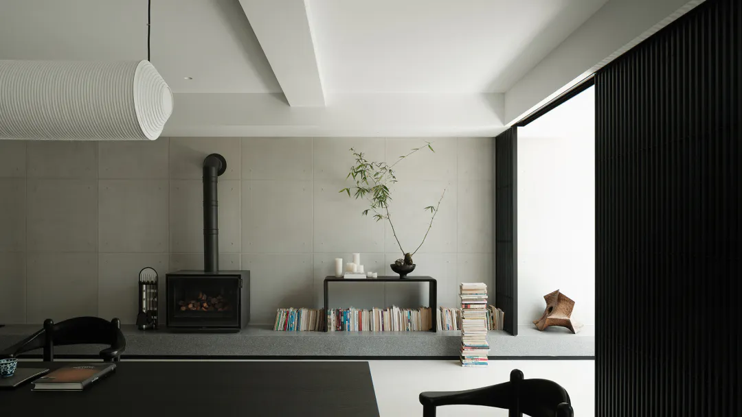 Modern minimalist living room with concrete walls, black fireplace, raised platform with books and bamboo plant, white cylindrical pendant lamp, and black dining chairs.