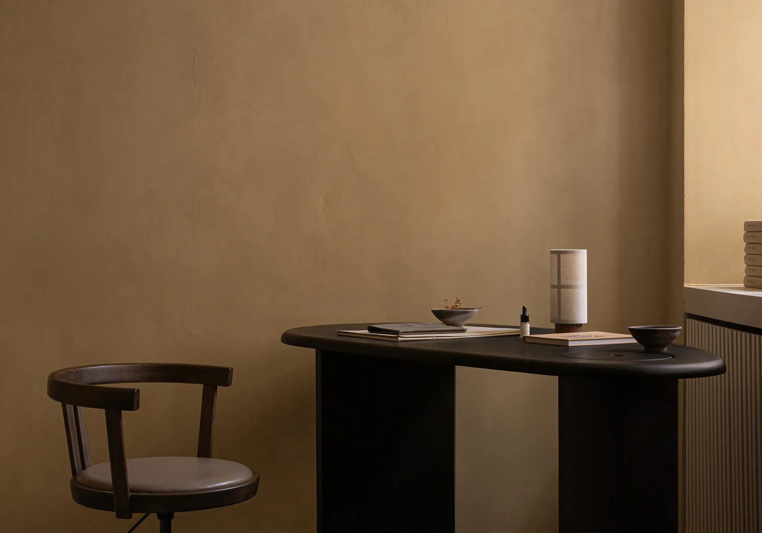 Minimalist home office corner with a dark walnut curved desk, wooden swivel chair with leather seat, ceramic bowls, table lamp, and books against a textured beige wall.