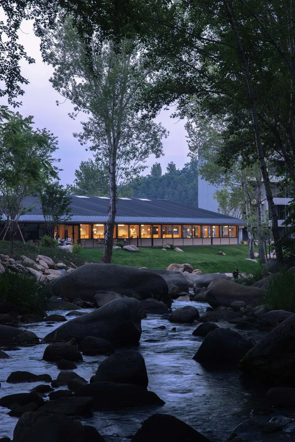 Modern concrete resort building with illuminated interior, situated next to a flowing stream with large boulders and surrounded by trees at twilight.