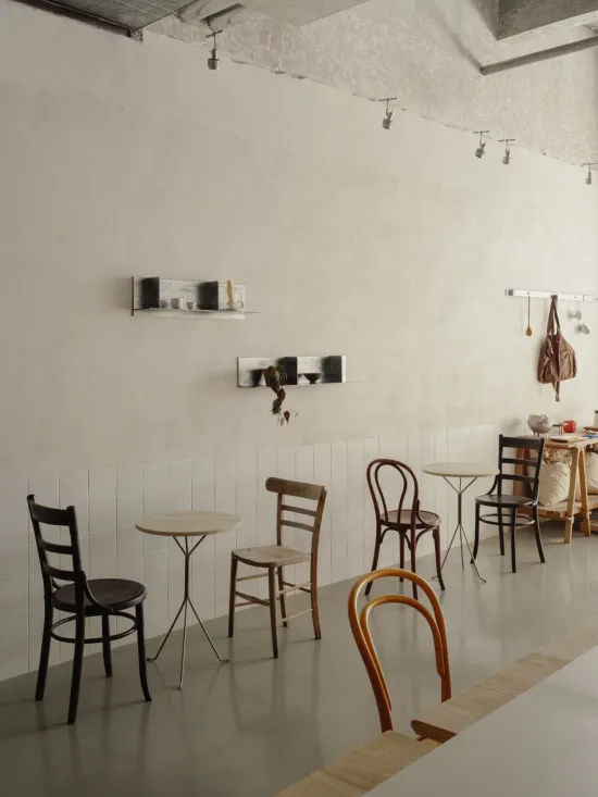 Bright minimalist cafe interior featuring a collection of wooden dining chairs including Thonet bentwood styles, small round tables, white tiled wainscoting, and textured plaster walls with floating shelves.