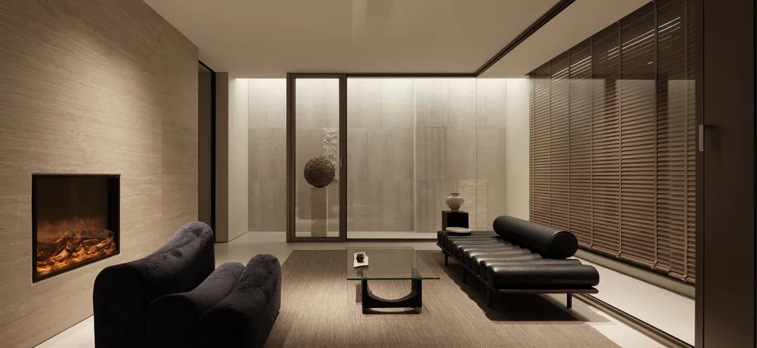 Modern minimalist living room featuring a travertine wall with built-in fireplace, dark velvet sofa, glass coffee table, and black leather chaise lounge against a wall of wooden blinds.