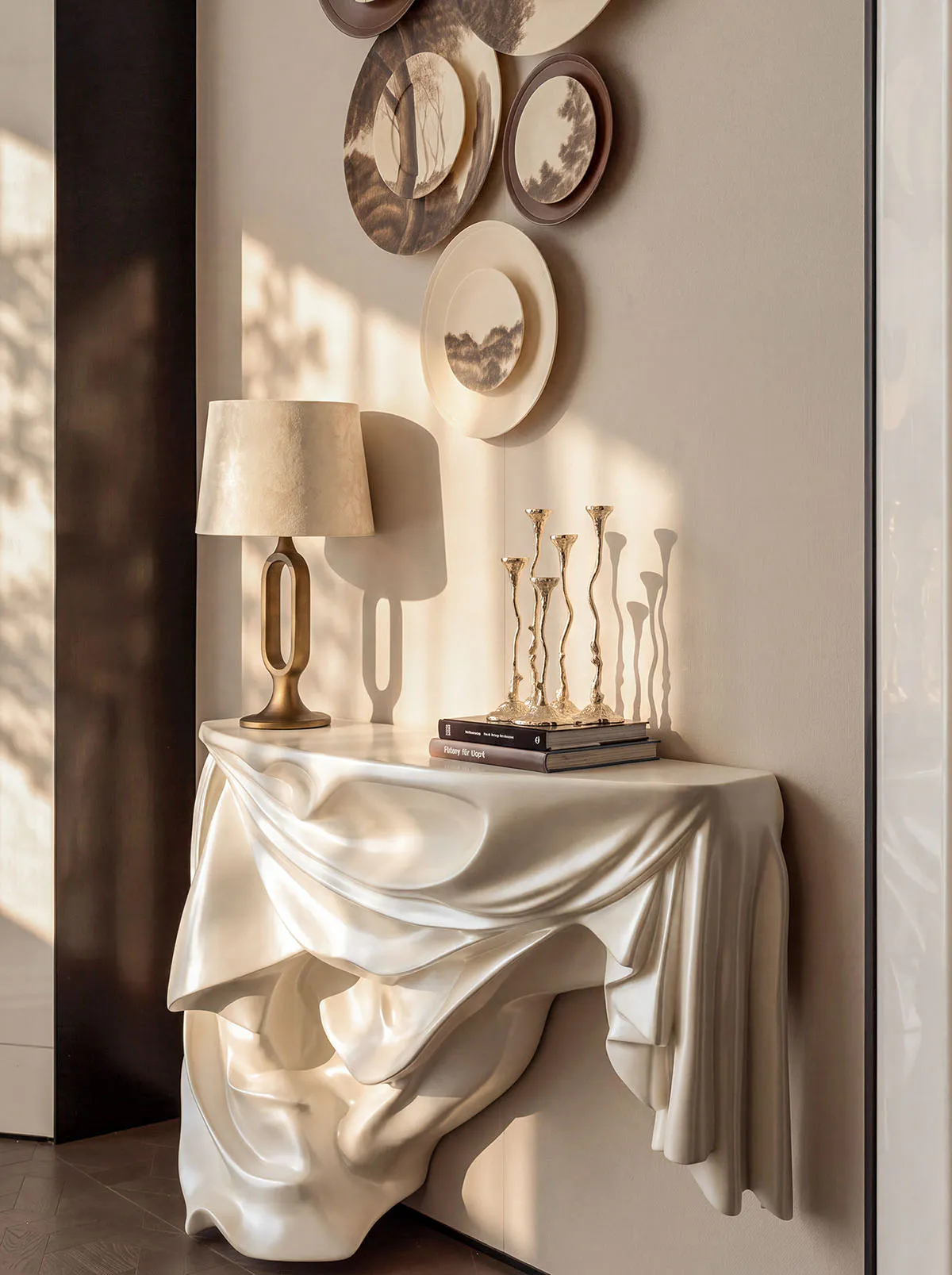 Modern sculptural console table with draped ivory finish, brass table lamp, and golden candlesticks against a neutral wall with decorative plates.