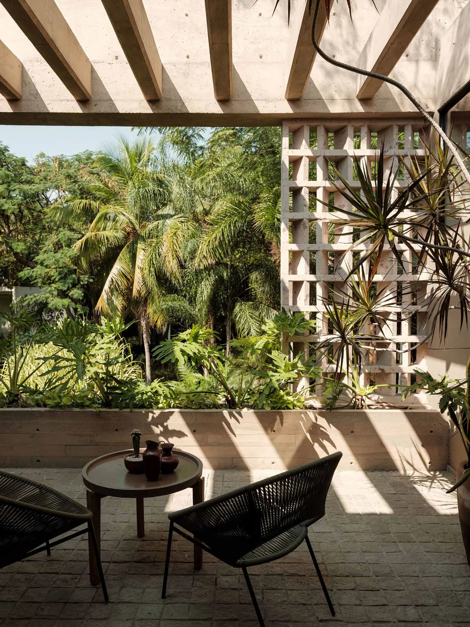 Modern tropical outdoor patio featuring concrete architecture, wooden pergola, dark woven lounge chairs, round wooden coffee table, and lush tropical plants under bright sunlight.