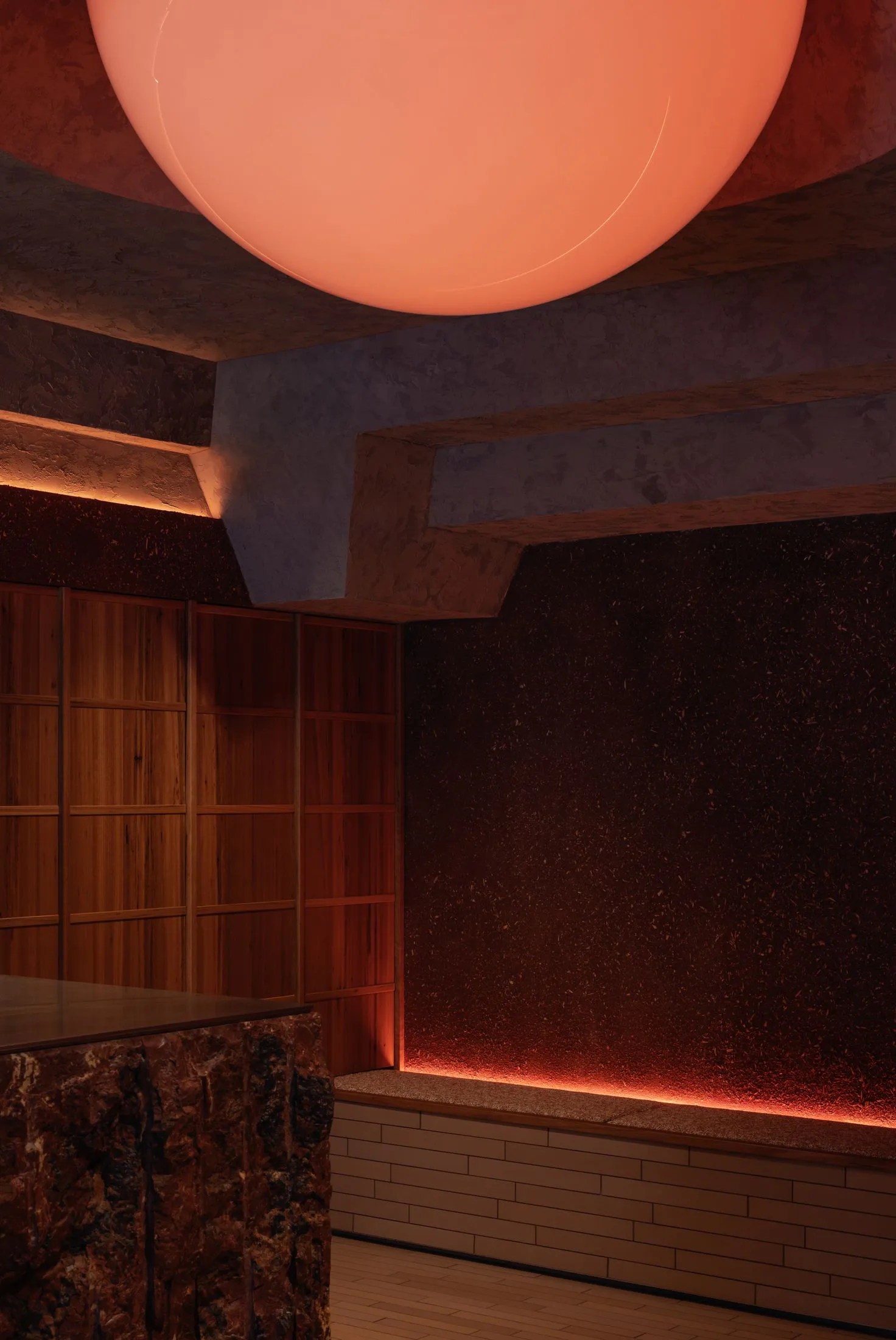 Interior view of a moody speakeasy-style bar featuring a large glowing red spherical pendant light, dark speckled terrazzo walls and counter, warm wood cabinetry, and layered architectural details with hidden lighting.
