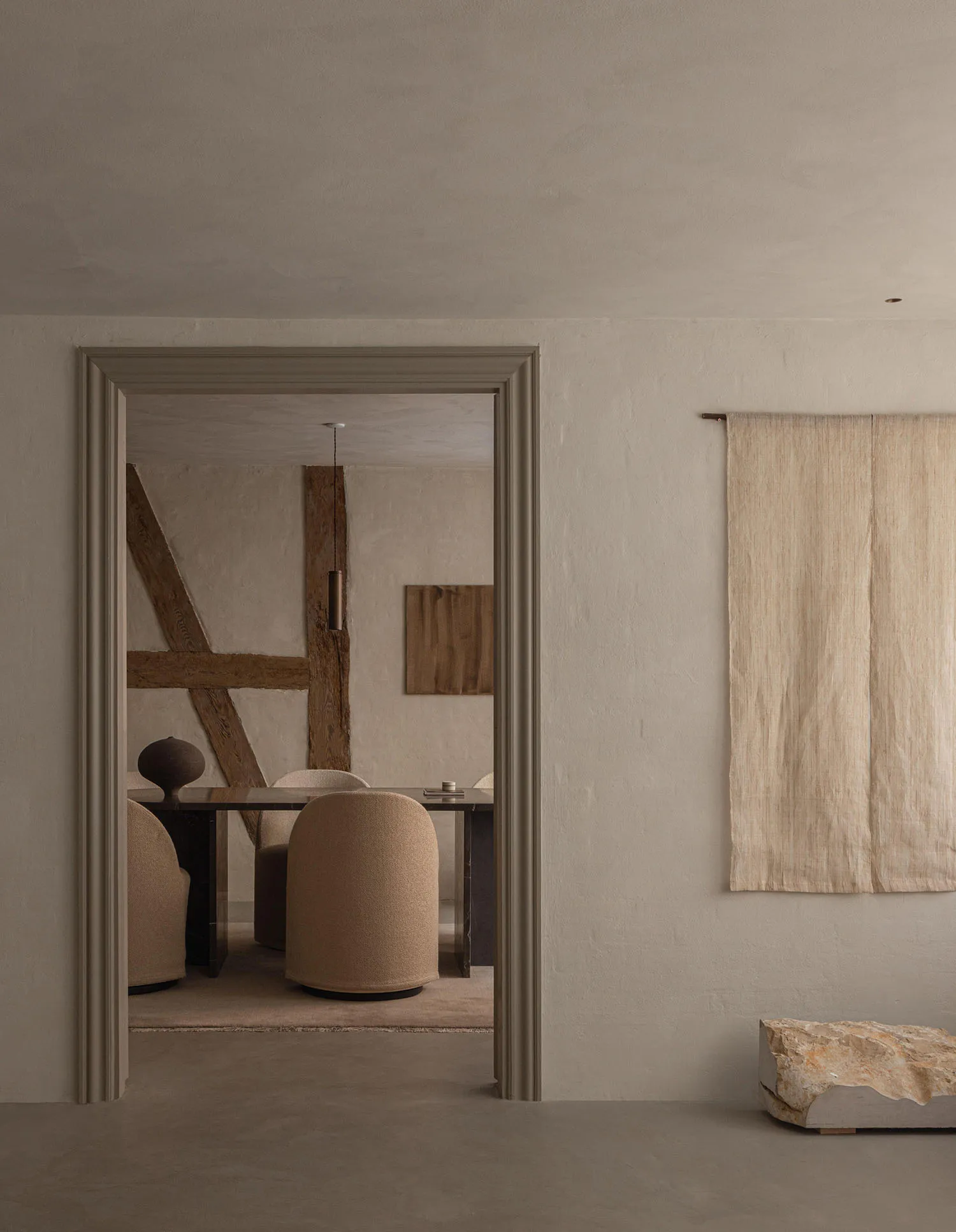 View from a neutral hallway into a wabi-sabi style dining room with beige boucle chairs, a dark stone table, exposed wooden beams, a hanging cylindrical pendant light, and a large raw travertine boulder on the floor in the foreground.