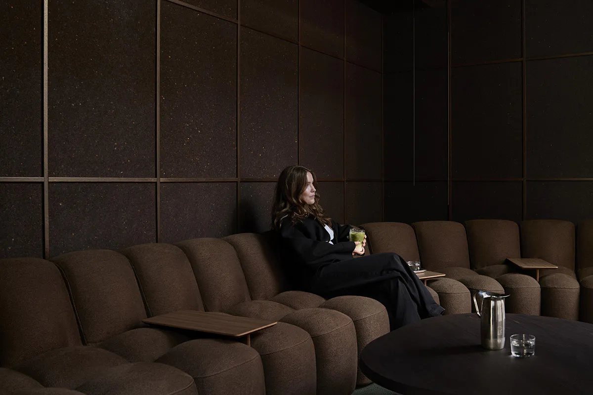 A woman with long brown hair wearing black sits on a brown modular sofa in a dark lounge, holding a glass of green matcha drink, surrounded by textured dark wall panels and a round coffee table with a metal pitcher.