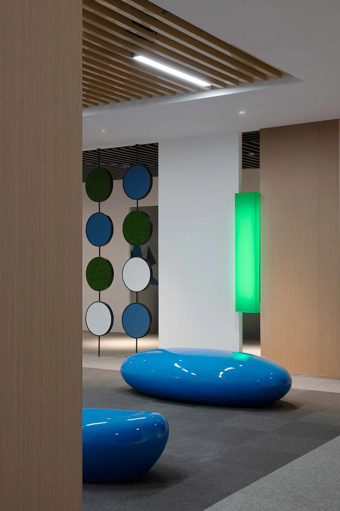 Modern corporate lounge interior with glossy blue organic-shaped seating pods, hanging circular moss and colored panels, wood slat ceiling, and a glowing green vertical light fixture.