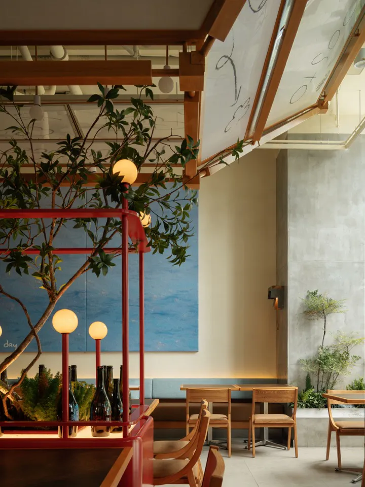 Modern restaurant interior featuring a vibrant red wooden trellis structure with integrated spherical pendant lights, a large blue ocean mural, wooden dining tables and chairs, and concrete walls with built-in banquette seating.