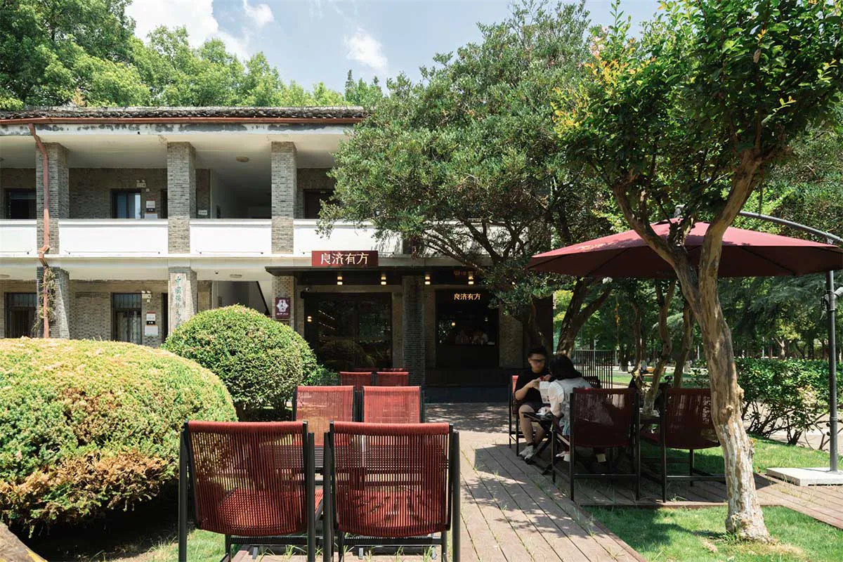 Outdoor seating area of a two-story brick heritage building converted into a cafe, featuring red mesh chairs on a wooden deck surrounded by lush greenery and large trees under a clear blue sky.