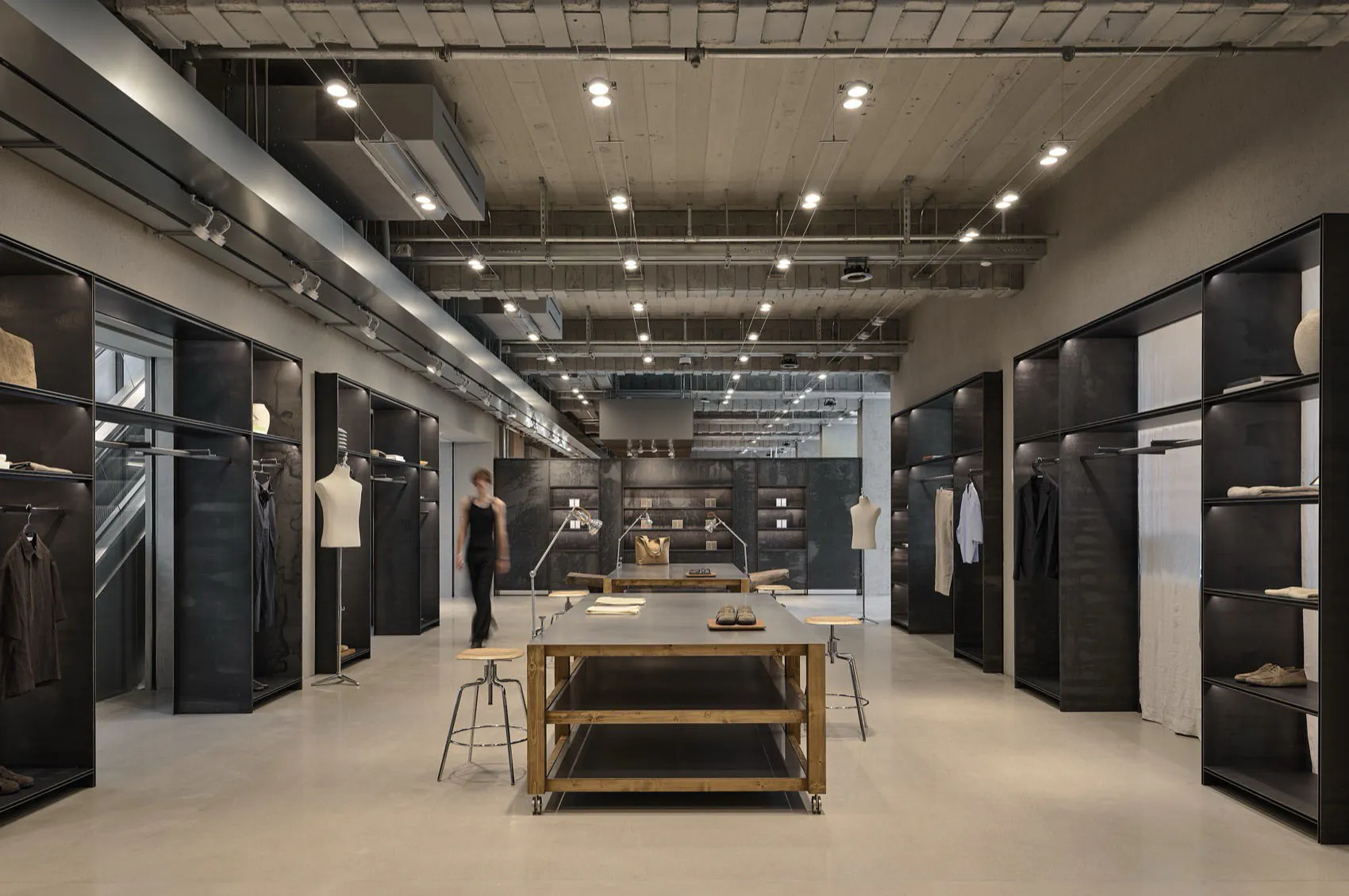 Modern industrial-style clothing boutique interior with raw concrete walls, black metal shelving, central wooden display table, and track lighting.