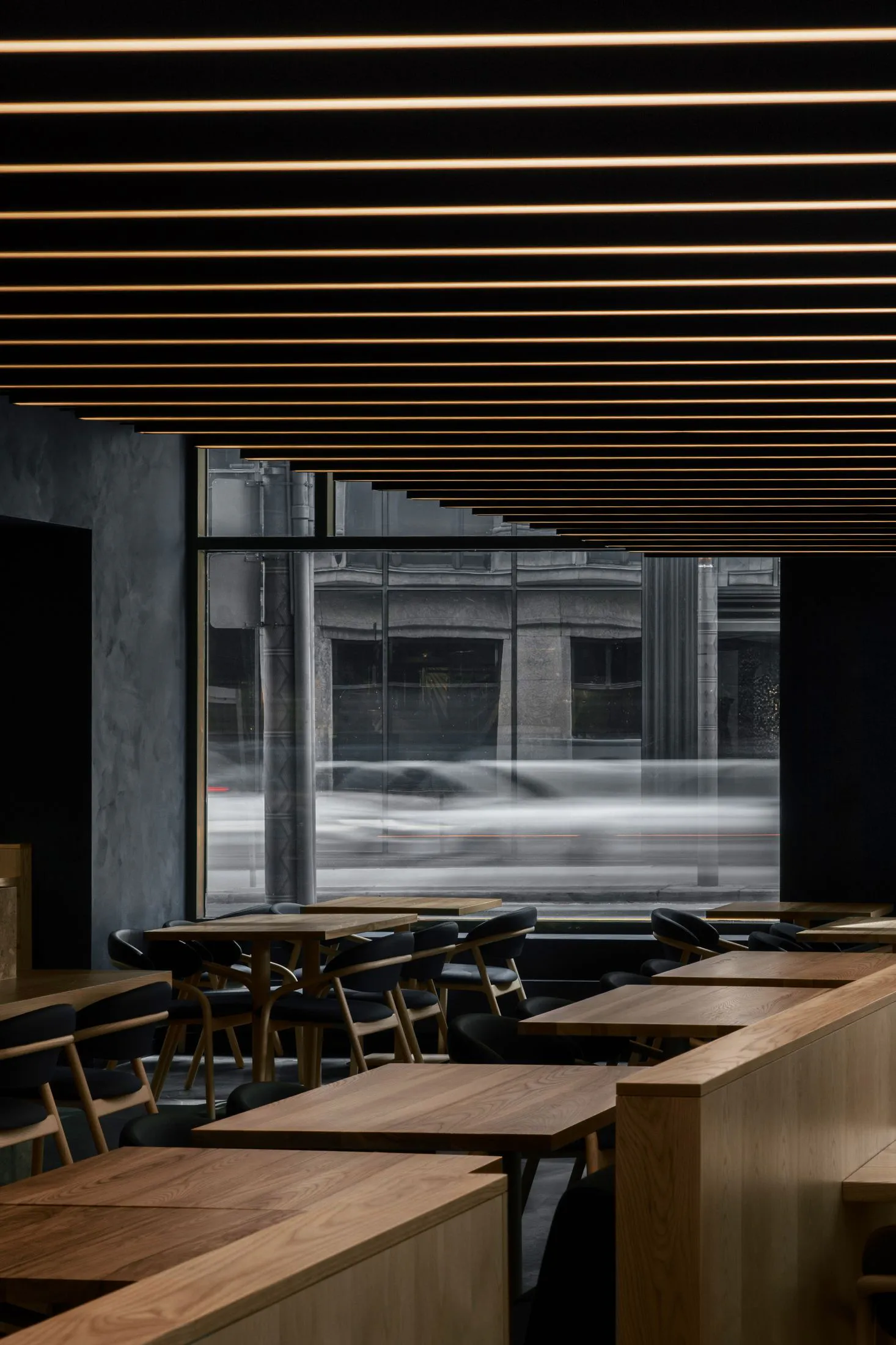 Modern minimalist restaurant interior featuring light oak wooden tables and benches, black upholstered chairs, dark concrete walls, and dramatic parallel linear LED lights on a black ceiling, with large windows showing motion-blurred city traffic outside.