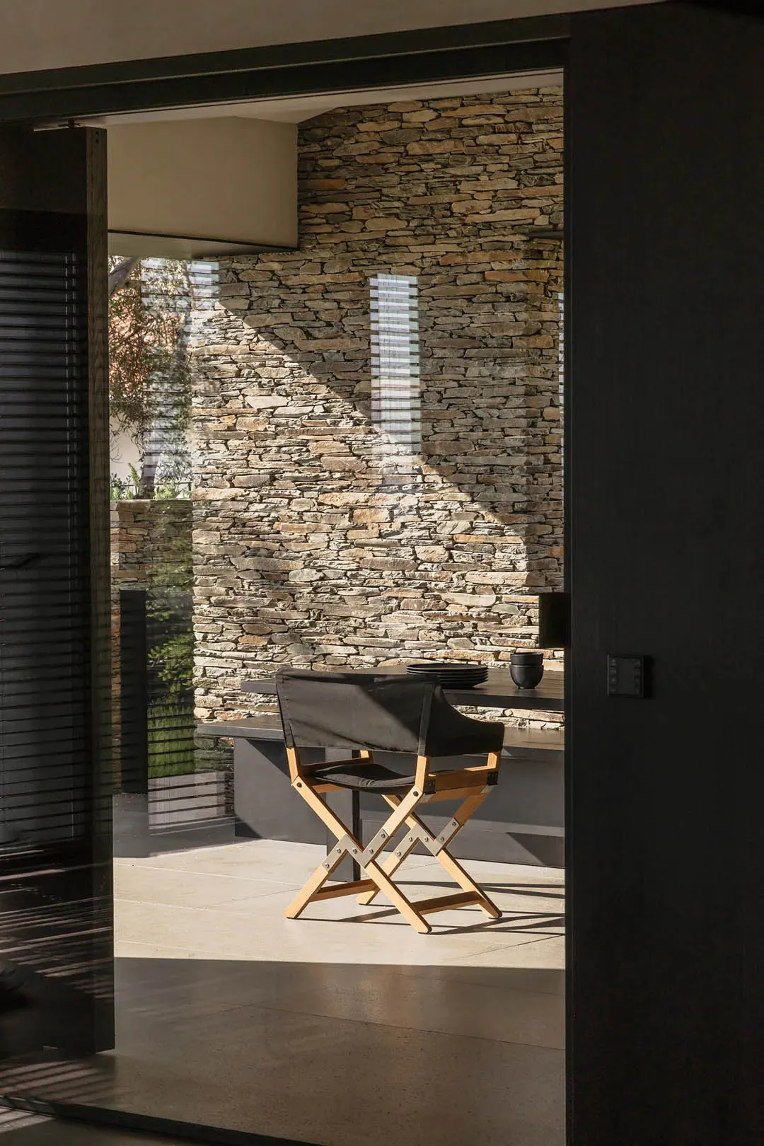 View through a dark modern doorway into a sunlit patio featuring a black canvas director’s chair with wooden frame, a dark counter with stacked plates and bowls, and a tall stacked stone wall with dramatic diagonal sunlight shadows.