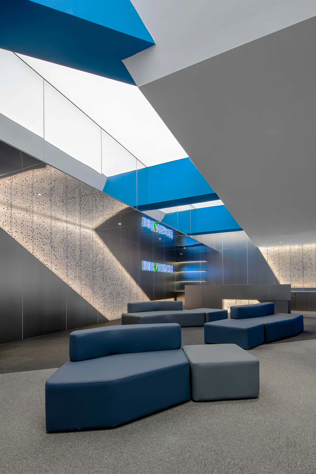 Contemporary tech company lounge with modular blue and grey sofas, dramatic angular blue ceiling panels, and large illuminated walls featuring intricate white circuit board patterns under a bright skylight.