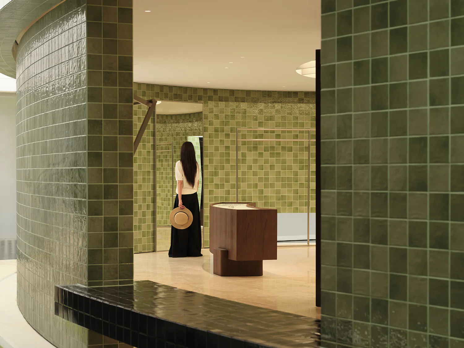 Woman standing in a modern boutique with curved walls covered in glossy green ceramic tiles and a minimalist walnut display counter.