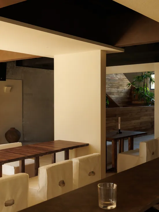 Interior view of a modern minimalist restaurant dining area with dark wood tables, off-white upholstered chairs, concrete walls, and warm ambient lighting.