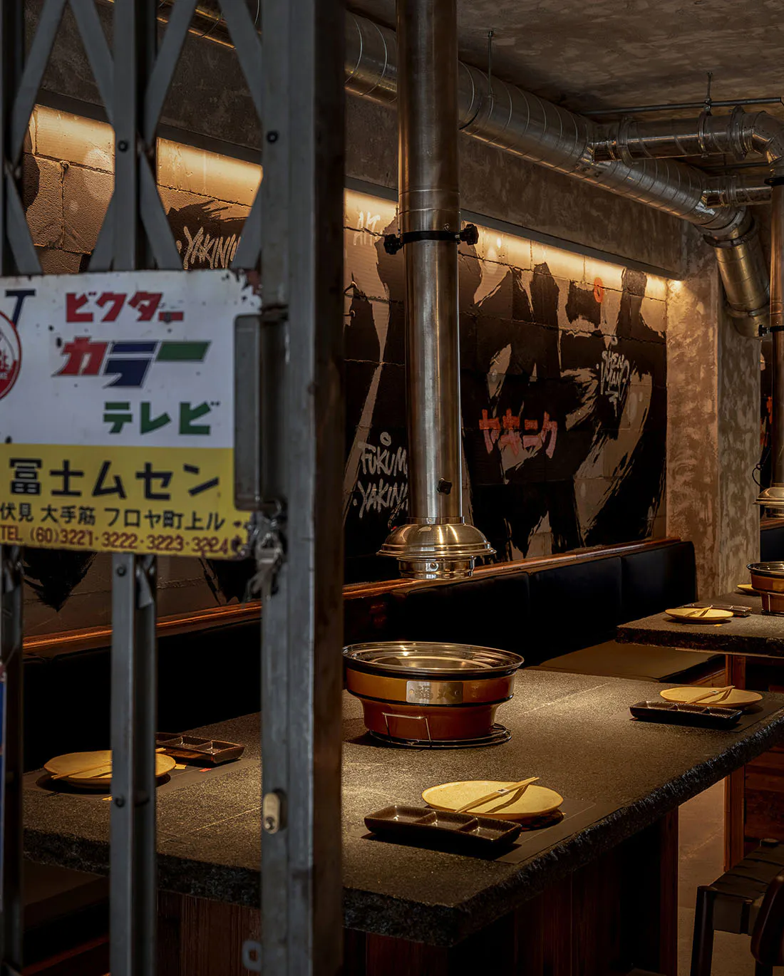 Interior view of a Japanese yakiniku restaurant featuring a dark granite counter, built-in copper hot pot, stainless steel ventilation hood, concrete walls with graffiti-style murals, and Japanese signage.