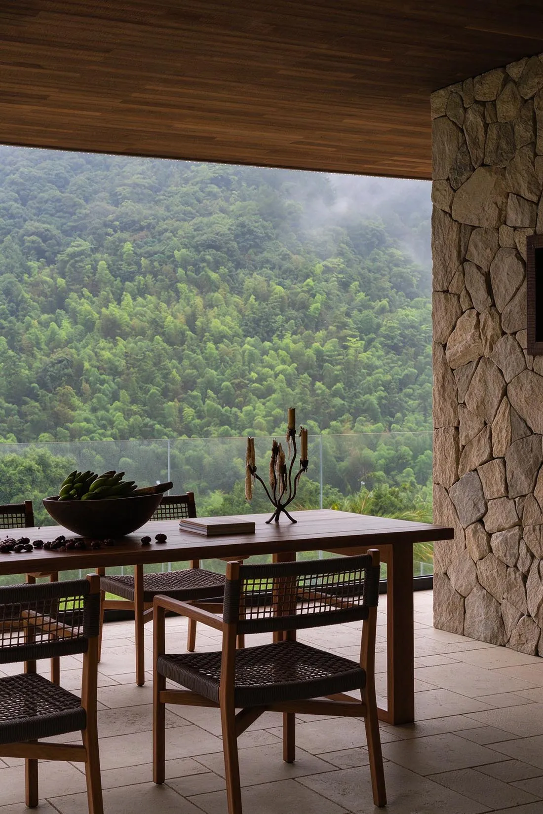 Outdoor dining table with woven chairs, large wooden bowl of green bananas, iron candelabra, set against a massive natural stone wall and expansive view of a lush green mountain covered in bamboo and mist.