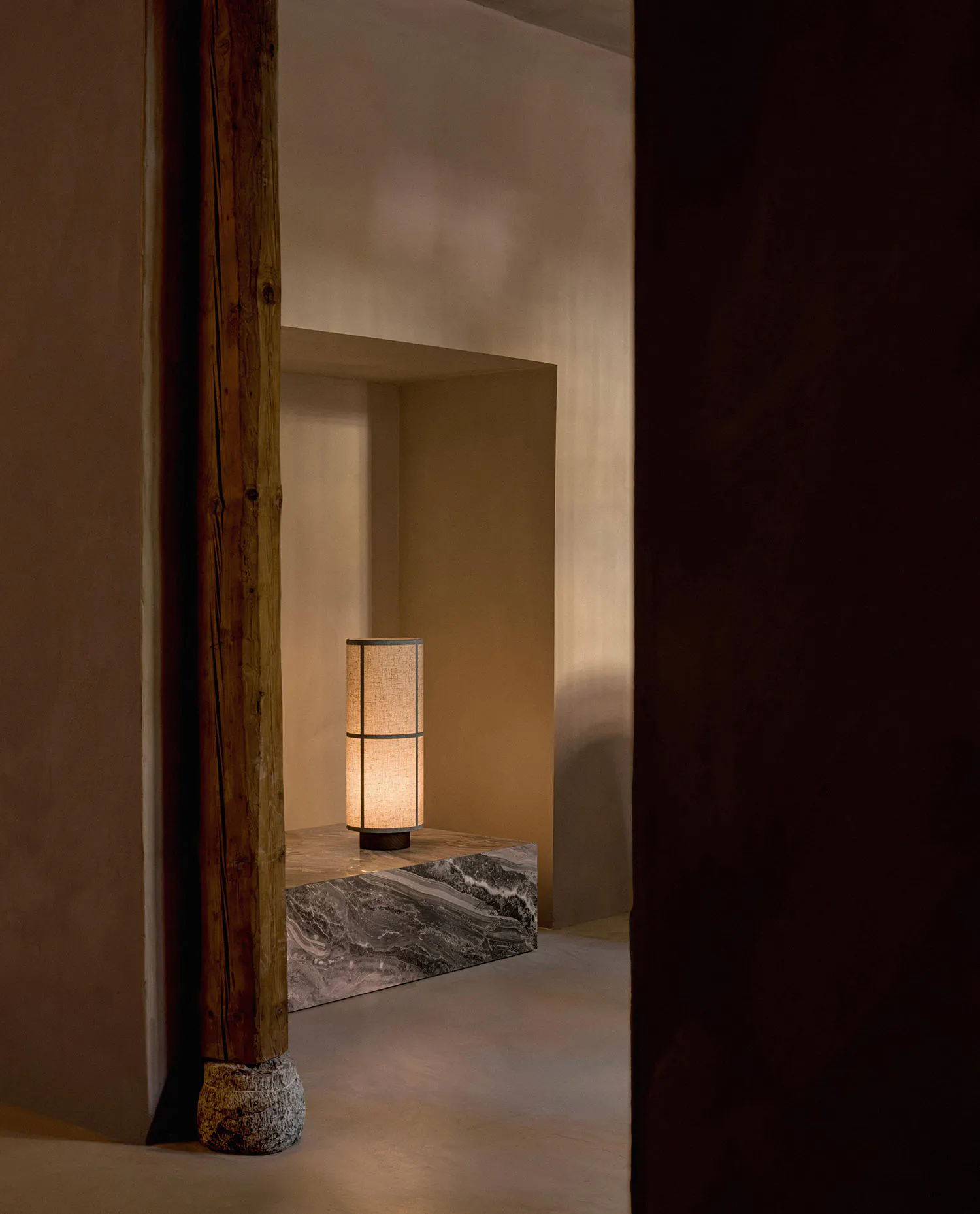 Warmly lit cylindrical table lamp placed on a grey marble ledge inside a recessed wall niche, beside a thick weathered wooden post with stone base in a minimalist interior.