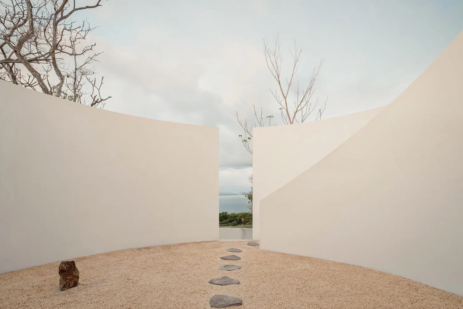 Minimalist outdoor courtyard with curved white concrete walls, gravel ground, irregular stone stepping path, a small dark rock sculpture, bare trees, and an opening revealing a sea view under a pale sky.