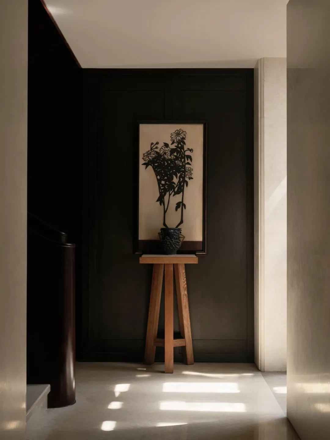 Modern dark entryway with light oak three-legged console table, dark ceramic vase, and large framed black chrysanthemum silhouette artwork on a dark wall.