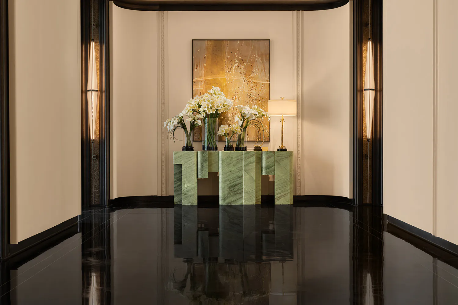 Modern luxury entryway featuring a green marble console table with white flower arrangements, large abstract gold painting, and dramatic vertical wall sconces reflected in a glossy black floor.