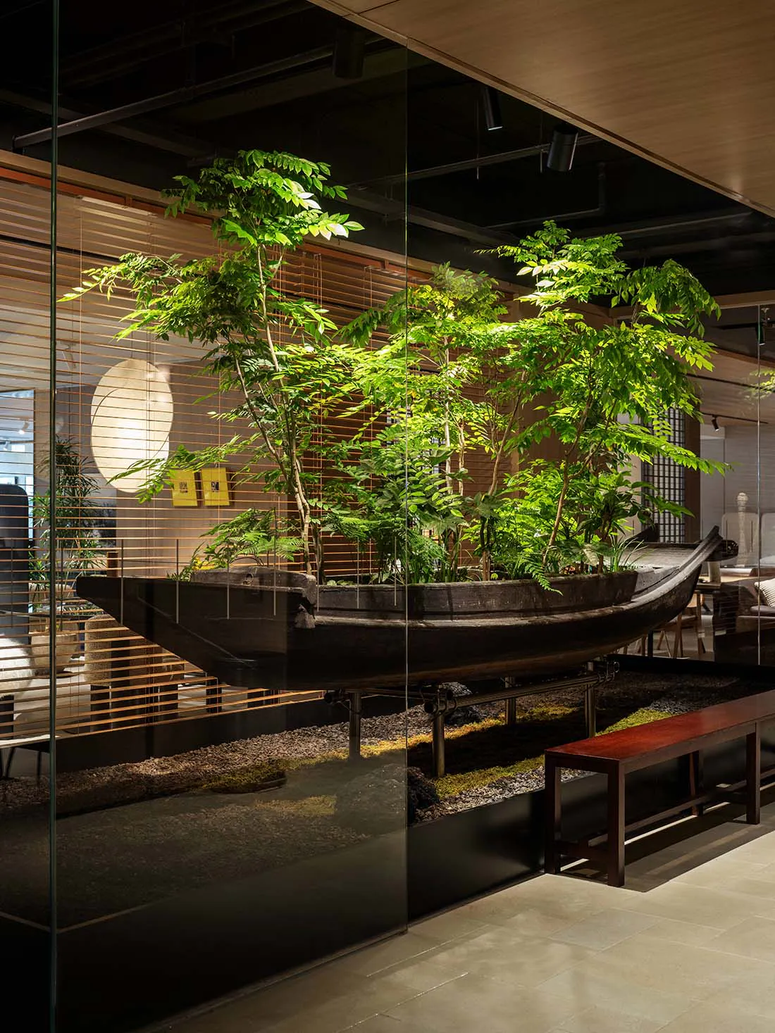 A large dark wooden boat filled with various green trees and plants is displayed as an art installation inside a modern living room, viewed through a glass wall with wooden blinds in the background.