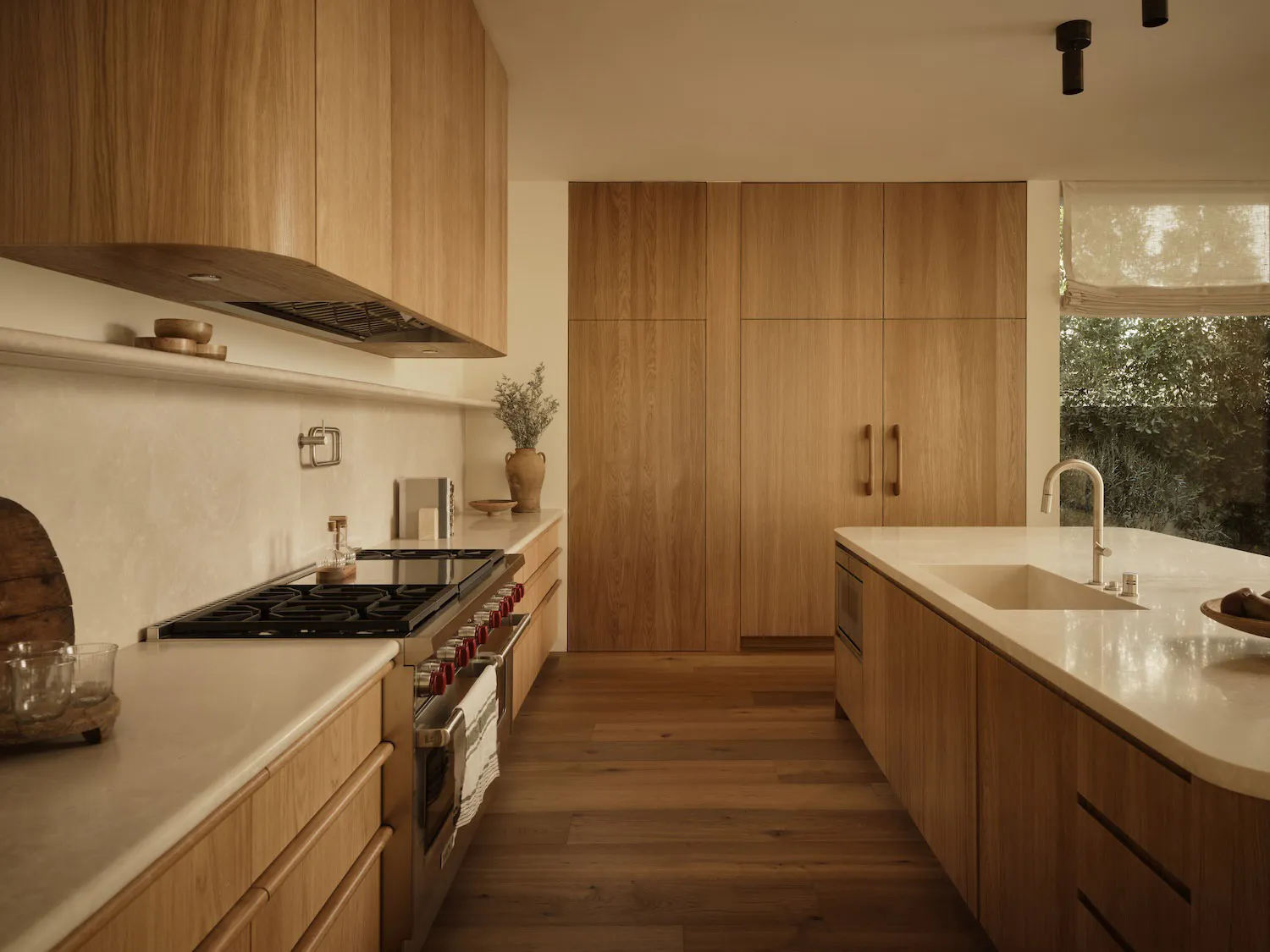 Modern minimalist kitchen featuring light oak cabinetry, white countertops, a central island with sink, and stainless steel range.